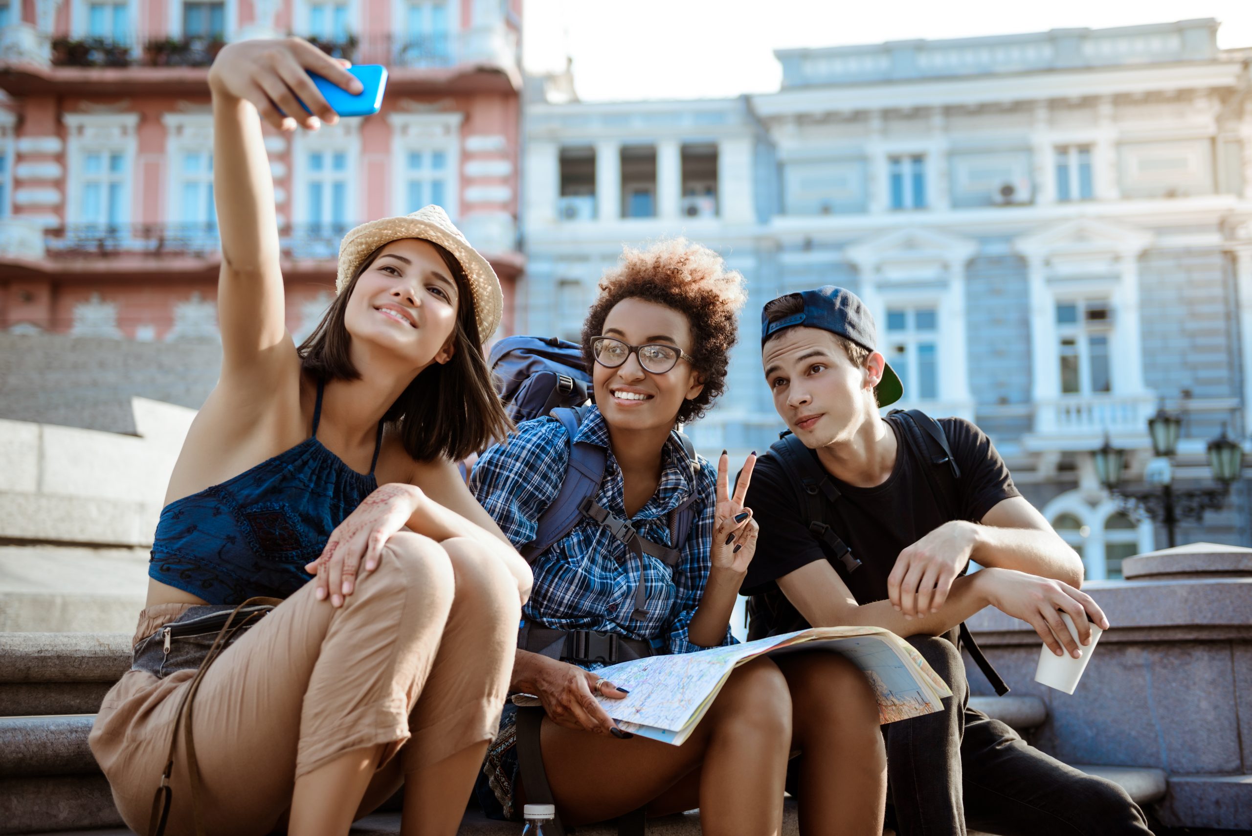 friends-travelers-with-backpacks-smiling-making-selfie-sitting-near-sight-scaled